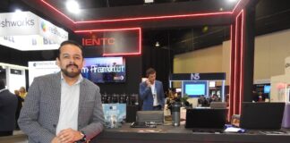 IENTC impulsa la conectividad empresarial para organizaciones que aceleran su transformación digital Fotografía en plano medio largo que captura a Rubén Vargas, manager del segmento corporativo de IENTC, posando de pie junto al módulo de atención de su empresa durante el encuentro de tecnología América Digital 2026. El directivo, con barba corta recortada, viste un saco gris claro con diseño de cuadros finos sobre una camisa formal clara y pantalón oscuro. Se apoya sutilmente en un mostrador negro decorado con tiras de luces LED de neón rojo y el logotipo de "IENTC TELECOMUNICACIONES". Sobre la mesa se aprecian folletos del "11° Congreso IA", computadoras portátiles y termos. Detrás de él, se observa a otro ejecutivo hablando por teléfono en un entorno de exposición dinámico con stands de las marcas freshworks y N5 bajo iluminación de plafón técnica.