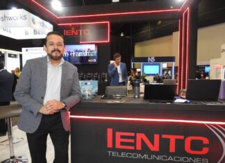 IENTC impulsa la conectividad empresarial para organizaciones que aceleran su transformación digital Fotografía en plano medio largo que captura a Rubén Vargas, manager del segmento corporativo de IENTC, posando de pie junto al módulo de atención de su empresa durante el encuentro de tecnología América Digital 2026. El directivo, con barba corta recortada, viste un saco gris claro con diseño de cuadros finos sobre una camisa formal clara y pantalón oscuro. Se apoya sutilmente en un mostrador negro decorado con tiras de luces LED de neón rojo y el logotipo de "IENTC TELECOMUNICACIONES". Sobre la mesa se aprecian folletos del "11° Congreso IA", computadoras portátiles y termos. Detrás de él, se observa a otro ejecutivo hablando por teléfono en un entorno de exposición dinámico con stands de las marcas freshworks y N5 bajo iluminación de plafón técnica.