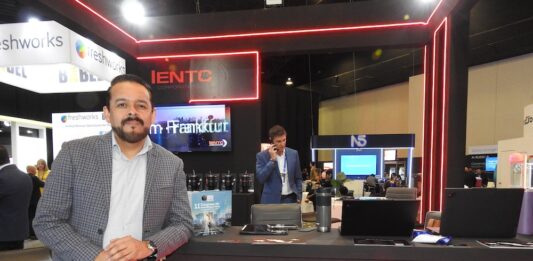 IENTC impulsa la conectividad empresarial para organizaciones que aceleran su transformación digital Fotografía en plano medio largo que captura a Rubén Vargas, manager del segmento corporativo de IENTC, posando de pie junto al módulo de atención de su empresa durante el encuentro de tecnología América Digital 2026. El directivo, con barba corta recortada, viste un saco gris claro con diseño de cuadros finos sobre una camisa formal clara y pantalón oscuro. Se apoya sutilmente en un mostrador negro decorado con tiras de luces LED de neón rojo y el logotipo de "IENTC TELECOMUNICACIONES". Sobre la mesa se aprecian folletos del "11° Congreso IA", computadoras portátiles y termos. Detrás de él, se observa a otro ejecutivo hablando por teléfono en un entorno de exposición dinámico con stands de las marcas freshworks y N5 bajo iluminación de plafón técnica.