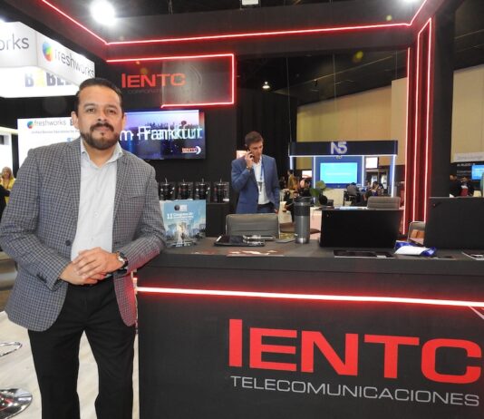 IENTC impulsa la conectividad empresarial para organizaciones que aceleran su transformación digital Fotografía en plano medio largo que captura a Rubén Vargas, manager del segmento corporativo de IENTC, posando de pie junto al módulo de atención de su empresa durante el encuentro de tecnología América Digital 2026. El directivo, con barba corta recortada, viste un saco gris claro con diseño de cuadros finos sobre una camisa formal clara y pantalón oscuro. Se apoya sutilmente en un mostrador negro decorado con tiras de luces LED de neón rojo y el logotipo de "IENTC TELECOMUNICACIONES". Sobre la mesa se aprecian folletos del "11° Congreso IA", computadoras portátiles y termos. Detrás de él, se observa a otro ejecutivo hablando por teléfono en un entorno de exposición dinámico con stands de las marcas freshworks y N5 bajo iluminación de plafón técnica.