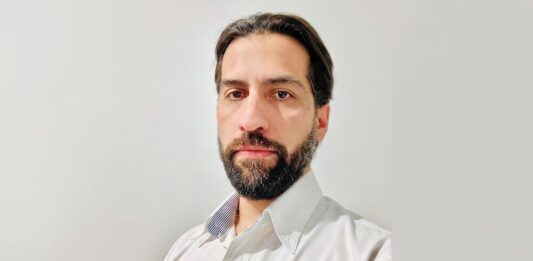 Eaton impulsa los UPS para infraestructura crítica con mayor disponibilidad, escalabilidad y eficiencia Fotografía de retrato en plano medio de Alejandro Lavín, Product Manager CPS de Eaton. El ejecutivo, de cabello oscuro, tez clara y barba, mira directamente a la cámara con una expresión neutral. Viste una camisa blanca de botones que lleva bordado el logotipo azul de la marca Eaton en el lado izquierdo del pecho, sobre un fondo liso de color gris claro.
