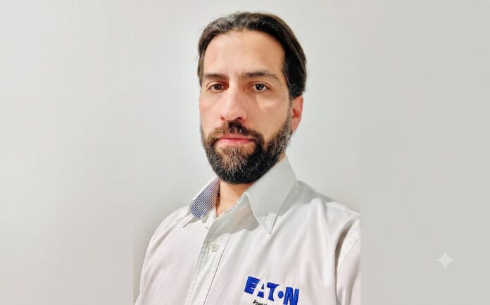 Fotografía de retrato en plano medio de Alejandro Lavín, Product Manager CPS de Eaton. El ejecutivo, de cabello oscuro, tez clara y barba, mira directamente a la cámara con una expresión neutral. Viste una camisa blanca de botones que lleva bordado el logotipo azul de la marca Eaton en el lado izquierdo del pecho, sobre un fondo liso de color gris claro.