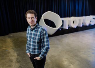 TOTVS fortalece su estrategia para el retail en América Latina con la integración de TOTVS Napse Fotografía de Marcelo Cosentino mirando a la cámara, de pie con las manos en los bolsillos y vistiendo una camisa a cuadros en tonos azules. En el fondo desenfocado, se aprecia un gran logotipo tridimensional de color blanco con la palabra TOTVS, situado sobre una plataforma negra frente a unas cortinas oscuras.
