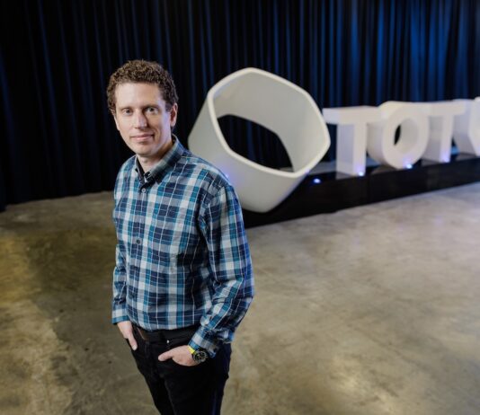TOTVS fortalece su estrategia para el retail en América Latina con la integración de TOTVS Napse Fotografía de Marcelo Cosentino mirando a la cámara, de pie con las manos en los bolsillos y vistiendo una camisa a cuadros en tonos azules. En el fondo desenfocado, se aprecia un gran logotipo tridimensional de color blanco con la palabra TOTVS, situado sobre una plataforma negra frente a unas cortinas oscuras.