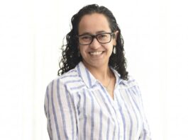 Sophos MDR acelera la respuesta a ciberamenazas con IA para fortalecer la resiliencia enterprise Retrato de estudio en plano medio y de frente de Daniela Arroyo, Channel Sales Engineer para LATAM en Sophos, posando sonriente sobre un fondo blanco uniforme. La especialista, de tez clara y cabello oscuro rizado de largo medio, usa anteojos de armazón negro, aretes colgantes plateados y una cadena delgada con un dije; viste una camisa blanca formal con líneas verticales azules y botones blancos.