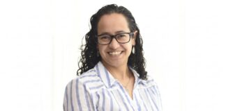 Sophos MDR acelera la respuesta a ciberamenazas con IA para fortalecer la resiliencia enterprise Retrato de estudio en plano medio y de frente de Daniela Arroyo, Channel Sales Engineer para LATAM en Sophos, posando sonriente sobre un fondo blanco uniforme. La especialista, de tez clara y cabello oscuro rizado de largo medio, usa anteojos de armazón negro, aretes colgantes plateados y una cadena delgada con un dije; viste una camisa blanca formal con líneas verticales azules y botones blancos.