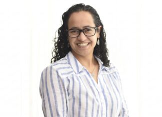 Sophos MDR acelera la respuesta a ciberamenazas con IA para fortalecer la resiliencia enterprise Retrato de estudio en plano medio y de frente de Daniela Arroyo, Channel Sales Engineer para LATAM en Sophos, posando sonriente sobre un fondo blanco uniforme. La especialista, de tez clara y cabello oscuro rizado de largo medio, usa anteojos de armazón negro, aretes colgantes plateados y una cadena delgada con un dije; viste una camisa blanca formal con líneas verticales azules y botones blancos.