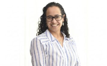 Sophos MDR acelera la respuesta a ciberamenazas con IA para fortalecer la resiliencia enterprise Retrato de estudio en plano medio y de frente de Daniela Arroyo, Channel Sales Engineer para LATAM en Sophos, posando sonriente sobre un fondo blanco uniforme. La especialista, de tez clara y cabello oscuro rizado de largo medio, usa anteojos de armazón negro, aretes colgantes plateados y una cadena delgada con un dije; viste una camisa blanca formal con líneas verticales azules y botones blancos.