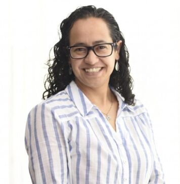 Sophos MDR acelera la respuesta a ciberamenazas con IA para fortalecer la resiliencia enterprise Retrato de estudio en plano medio y de frente de Daniela Arroyo, Channel Sales Engineer para LATAM en Sophos, posando sonriente sobre un fondo blanco uniforme. La especialista, de tez clara y cabello oscuro rizado de largo medio, usa anteojos de armazón negro, aretes colgantes plateados y una cadena delgada con un dije; viste una camisa blanca formal con líneas verticales azules y botones blancos.