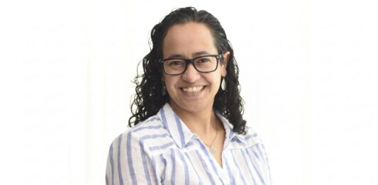 Sophos MDR acelera la respuesta a ciberamenazas con IA para fortalecer la resiliencia enterprise Retrato de estudio en plano medio y de frente de Daniela Arroyo, Channel Sales Engineer para LATAM en Sophos, posando sonriente sobre un fondo blanco uniforme. La especialista, de tez clara y cabello oscuro rizado de largo medio, usa anteojos de armazón negro, aretes colgantes plateados y una cadena delgada con un dije; viste una camisa blanca formal con líneas verticales azules y botones blancos.