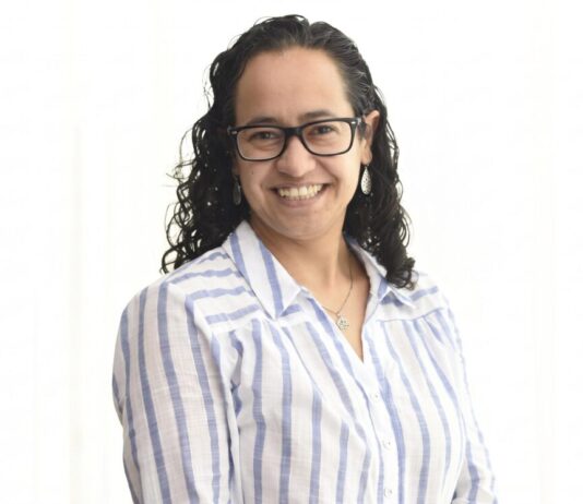 Sophos MDR acelera la respuesta a ciberamenazas con IA para fortalecer la resiliencia enterprise Retrato de estudio en plano medio y de frente de Daniela Arroyo, Channel Sales Engineer para LATAM en Sophos, posando sonriente sobre un fondo blanco uniforme. La especialista, de tez clara y cabello oscuro rizado de largo medio, usa anteojos de armazón negro, aretes colgantes plateados y una cadena delgada con un dije; viste una camisa blanca formal con líneas verticales azules y botones blancos.