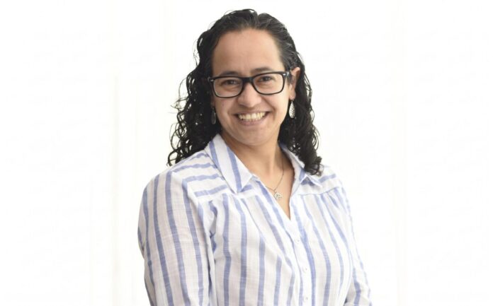 Daniela Arroyo analiza los indicadores de respuesta inmediata frente a incidentes en grandes corporativos Retrato de estudio en plano medio y de frente de Daniela Arroyo, Channel Sales Engineer para LATAM en Sophos, posando sonriente sobre un fondo blanco uniforme. La especialista, de tez clara y cabello oscuro rizado de largo medio, usa anteojos de armazón negro, aretes colgantes plateados y una cadena delgada con un dije; viste una camisa blanca formal con líneas verticales azules y botones blancos.