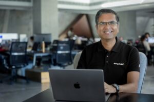 Fotografía de Rajesh Ganesan sonriendo cálidamente hacia la lente. Es un hombre con gafas que viste una playera tipo polo color negro con el logotipo de ManageEngine. Se encuentra sentado trabajando frente a una computadora portátil gris sobre un escritorio oscuro, dentro de una oficina moderna, amplia y con buena iluminación natural.