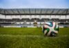 Evertec destaca la infraestructura de pagos como pieza clave para el Mundial 2026 Fotografía de un balón de fútbol sobre el césped verde de una cancha, con las gradas y la estructura de un gran estadio vacante desenfocadas en el fondo bajo un cielo nublado.