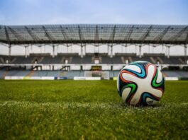 Evertec destaca la infraestructura de pagos como pieza clave para el Mundial 2026 Fotografía de un balón de fútbol sobre el césped verde de una cancha, con las gradas y la estructura de un gran estadio vacante desenfocadas en el fondo bajo un cielo nublado.