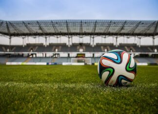 Evertec destaca la infraestructura de pagos como pieza clave para el Mundial 2026 Fotografía de un balón de fútbol sobre el césped verde de una cancha, con las gradas y la estructura de un gran estadio vacante desenfocadas en el fondo bajo un cielo nublado.