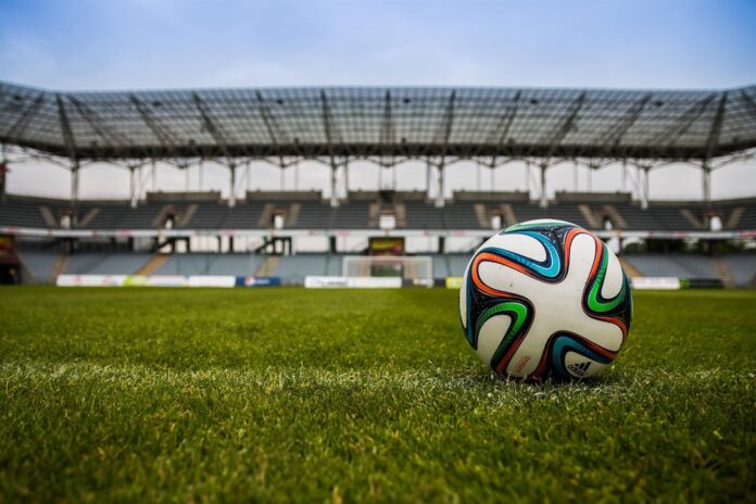 Fotografía de un balón de fútbol sobre el césped verde de una cancha, con las gradas y la estructura de un gran estadio vacante desenfocadas en el fondo bajo un cielo nublado.