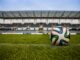 Evertec destaca la infraestructura de pagos como pieza clave para el Mundial 2026 Fotografía de un balón de fútbol sobre el césped verde de una cancha, con las gradas y la estructura de un gran estadio vacante desenfocadas en el fondo bajo un cielo nublado.