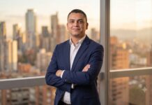 Servinformación convierte los datos en ventaja competitiva con soluciones de geolocalización inteligente Fotografía de Kevin Alfonso, vistiendo un traje azul oscuro sobre una camisa blanca, posando de pie con los brazos cruzados. Se encuentra frente a un amplio ventanal que ofrece una vista panorámica de una ciudad iluminada por la luz dorada del atardecer.