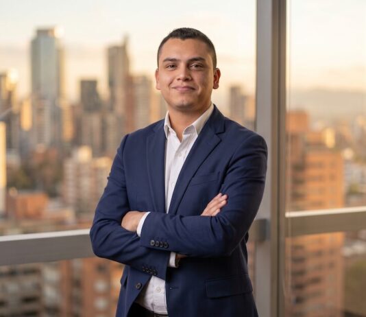 Servinformación convierte los datos en ventaja competitiva con soluciones de geolocalización inteligente Fotografía de Kevin Alfonso, vistiendo un traje azul oscuro sobre una camisa blanca, posando de pie con los brazos cruzados. Se encuentra frente a un amplio ventanal que ofrece una vista panorámica de una ciudad iluminada por la luz dorada del atardecer.