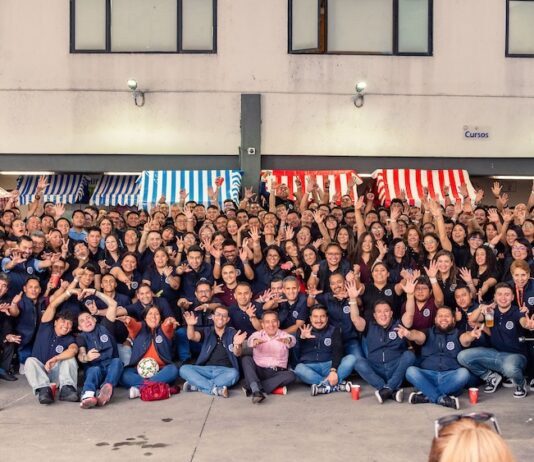 Tecnosinergia redefine la seguridad Enterprise con ecosistemas inteligentes Fotografía en plano general amplio que muestra a todo el ecosistema de colaboradores de Tecnosinergia posando juntos al aire libre frente al edificio corporativo. Más de cien personas, la mayoría vistiendo el chaleco azul institucional, posan sentadas, de rodillas y de pie, saludando y sonriendo a la cámara con las manos en alto, formando señas de amor y paz y gestos festivos. A los costados y en la parte trasera se alcanzan a ver estructuras de puestos de feria de color rojo y azul, mientras que a la derecha en primer plano destaca parcialmente una mesa de futbolito azul del Real Madrid vs. Barcelona.