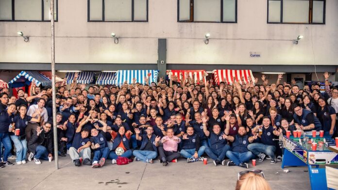 Fotografía en plano general amplio que muestra a todo el ecosistema de colaboradores de Tecnosinergia posando juntos al aire libre frente al edificio corporativo. Más de cien personas, la mayoría vistiendo el chaleco azul institucional, posan sentadas, de rodillas y de pie, saludando y sonriendo a la cámara con las manos en alto, formando señas de amor y paz y gestos festivos. A los costados y en la parte trasera se alcanzan a ver estructuras de puestos de feria de color rojo y azul, mientras que a la derecha en primer plano destaca parcialmente una mesa de futbolito azul del Real Madrid vs. Barcelona.