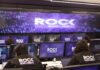KIO IT Services lanza The ROCK para reforzar la continuidad de los negocios frente a ciberataques impulsados por IA Una fotografía que muestra una sala de operaciones de ciberseguridad con varias filas de estaciones de trabajo, donde analistas de seguridad están de espaldas, monitoreando múltiples pantallas. Cada monitor individual y la pantalla gigante de pared al fondo muestran el logotipo "The ROCK SOC by KIO IT SERVICES" en letras blancas sobre un fondo azul oscuro con puntos de luz. Dos analistas en primer plano llevan chalecos negros con el logotipo bordado "The ROCK SOC by KIO IT SERVICES" en la espalda.
