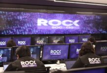 KIO IT Services lanza The ROCK para reforzar la continuidad de los negocios frente a ciberataques impulsados por IA Una fotografía que muestra una sala de operaciones de ciberseguridad con varias filas de estaciones de trabajo, donde analistas de seguridad están de espaldas, monitoreando múltiples pantallas. Cada monitor individual y la pantalla gigante de pared al fondo muestran el logotipo "The ROCK SOC by KIO IT SERVICES" en letras blancas sobre un fondo azul oscuro con puntos de luz. Dos analistas en primer plano llevan chalecos negros con el logotipo bordado "The ROCK SOC by KIO IT SERVICES" en la espalda.