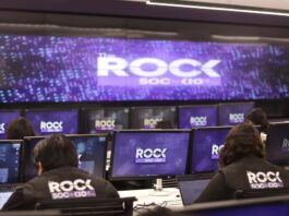 KIO IT Services lanza The ROCK para reforzar la continuidad de los negocios frente a ciberataques impulsados por IA Una fotografía que muestra una sala de operaciones de ciberseguridad con varias filas de estaciones de trabajo, donde analistas de seguridad están de espaldas, monitoreando múltiples pantallas. Cada monitor individual y la pantalla gigante de pared al fondo muestran el logotipo "The ROCK SOC by KIO IT SERVICES" en letras blancas sobre un fondo azul oscuro con puntos de luz. Dos analistas en primer plano llevan chalecos negros con el logotipo bordado "The ROCK SOC by KIO IT SERVICES" en la espalda.