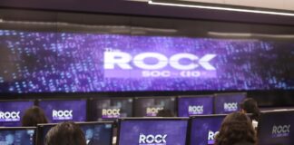 KIO IT Services lanza The ROCK para reforzar la continuidad de los negocios frente a ciberataques impulsados por IA Una fotografía que muestra una sala de operaciones de ciberseguridad con varias filas de estaciones de trabajo, donde analistas de seguridad están de espaldas, monitoreando múltiples pantallas. Cada monitor individual y la pantalla gigante de pared al fondo muestran el logotipo "The ROCK SOC by KIO IT SERVICES" en letras blancas sobre un fondo azul oscuro con puntos de luz. Dos analistas en primer plano llevan chalecos negros con el logotipo bordado "The ROCK SOC by KIO IT SERVICES" en la espalda.