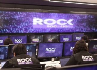 KIO IT Services lanza The ROCK para reforzar la continuidad de los negocios frente a ciberataques impulsados por IA Una fotografía que muestra una sala de operaciones de ciberseguridad con varias filas de estaciones de trabajo, donde analistas de seguridad están de espaldas, monitoreando múltiples pantallas. Cada monitor individual y la pantalla gigante de pared al fondo muestran el logotipo "The ROCK SOC by KIO IT SERVICES" en letras blancas sobre un fondo azul oscuro con puntos de luz. Dos analistas en primer plano llevan chalecos negros con el logotipo bordado "The ROCK SOC by KIO IT SERVICES" en la espalda.
