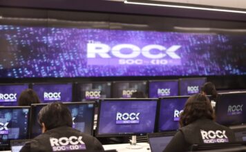 KIO IT Services lanza The ROCK para reforzar la continuidad de los negocios frente a ciberataques impulsados por IA Una fotografía que muestra una sala de operaciones de ciberseguridad con varias filas de estaciones de trabajo, donde analistas de seguridad están de espaldas, monitoreando múltiples pantallas. Cada monitor individual y la pantalla gigante de pared al fondo muestran el logotipo "The ROCK SOC by KIO IT SERVICES" en letras blancas sobre un fondo azul oscuro con puntos de luz. Dos analistas en primer plano llevan chalecos negros con el logotipo bordado "The ROCK SOC by KIO IT SERVICES" en la espalda.