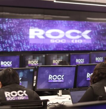 KIO IT Services lanza The ROCK para reforzar la continuidad de los negocios frente a ciberataques impulsados por IA Una fotografía que muestra una sala de operaciones de ciberseguridad con varias filas de estaciones de trabajo, donde analistas de seguridad están de espaldas, monitoreando múltiples pantallas. Cada monitor individual y la pantalla gigante de pared al fondo muestran el logotipo "The ROCK SOC by KIO IT SERVICES" en letras blancas sobre un fondo azul oscuro con puntos de luz. Dos analistas en primer plano llevan chalecos negros con el logotipo bordado "The ROCK SOC by KIO IT SERVICES" en la espalda.