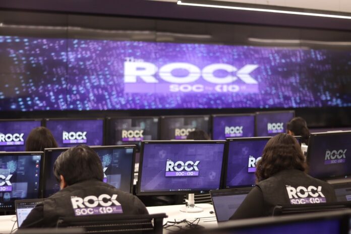 Inauguración de The ROCK, el nuevo modelo operativo de ciberseguridad de KIO IT Services diseñado para enfrentar amenazas automatizadas Una fotografía que muestra una sala de operaciones de ciberseguridad con varias filas de estaciones de trabajo, donde analistas de seguridad están de espaldas, monitoreando múltiples pantallas. Cada monitor individual y la pantalla gigante de pared al fondo muestran el logotipo