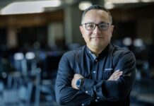 ManageEngine revela que las empresas mexicanas apuestan por consolidar TI para acelerar el valor de la inteligencia artificial Fotografía de medio cuerpo de un hombre, Wilson Calderón, que mira a la cámara con una expresión serena y profesional. Usa anteojos oscuros y una camisa negra de manga larga con el logotipo de ManageEngine bordado en el pecho. Mantiene los brazos cruzados y se encuentra de pie frente a un entorno de oficina que aparece completamente desenfocado en el fondo.