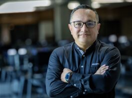 ManageEngine revela que las empresas mexicanas apuestan por consolidar TI para acelerar el valor de la inteligencia artificial Fotografía de medio cuerpo de un hombre, Wilson Calderón, que mira a la cámara con una expresión serena y profesional. Usa anteojos oscuros y una camisa negra de manga larga con el logotipo de ManageEngine bordado en el pecho. Mantiene los brazos cruzados y se encuentra de pie frente a un entorno de oficina que aparece completamente desenfocado en el fondo.