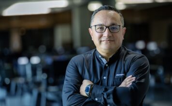 ManageEngine revela que las empresas mexicanas apuestan por consolidar TI para acelerar el valor de la inteligencia artificial Fotografía de medio cuerpo de un hombre, Wilson Calderón, que mira a la cámara con una expresión serena y profesional. Usa anteojos oscuros y una camisa negra de manga larga con el logotipo de ManageEngine bordado en el pecho. Mantiene los brazos cruzados y se encuentra de pie frente a un entorno de oficina que aparece completamente desenfocado en el fondo.