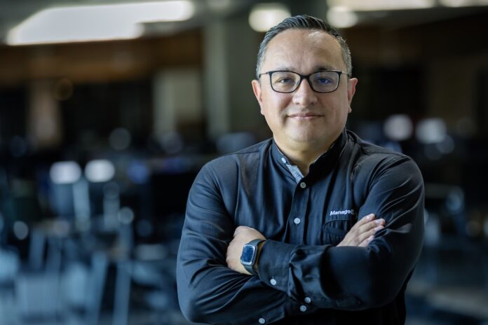 La consolidación tecnológica como la nueva ventaja competitiva en México Fotografía de medio cuerpo de un hombre, Wilson Calderón, que mira a la cámara con una expresión serena y profesional. Usa anteojos oscuros y una camisa negra de manga larga con el logotipo de ManageEngine bordado en el pecho. Mantiene los brazos cruzados y se encuentra de pie frente a un entorno de oficina que aparece completamente desenfocado en el fondo.