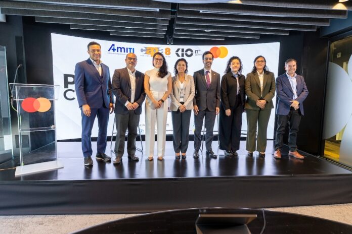 Fotografía frontal de un grupo de ocho personas, conformado por hombres y mujeres en vestimenta formal de negocios, posando de pie y sonriendo sobre un escenario. A la izquierda del grupo hay un podio transparente que muestra el logotipo de Mastercard. En la parte trasera, una gran pantalla curva iluminada exhibe parte del texto del evento y los logotipos oficiales de AMITI, Capa 8, KIO IT Services y Mastercard.