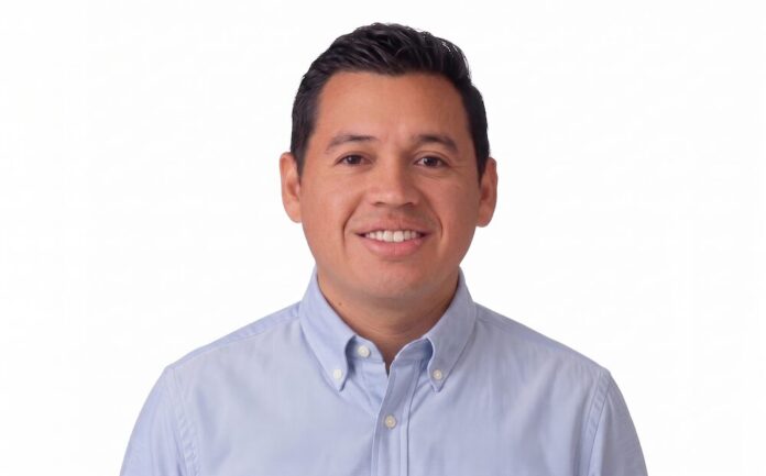 Fotografía de retrato de Xavier Arroyo, hombre de tez clara, cabello oscuro y corto, sonriendo directamente a la cámara. Viste una camisa de botones en tono azul claro y se encuentra sobre un fondo completamente blanco.