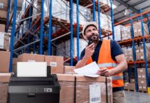 Epson mantiene vigente la impresión por matriz de puntos en operaciones críticas Composición visual que muestra en primer plano una impresora de matriz de puntos Epson de color oscuro con papel continuo instalado. En el fondo, desenfocado para dar profundidad, se observa a un trabajador de logística con casco blanco y chaleco de seguridad naranja, revisando documentos físicos y comunicándose por radio frente a estantes industriales llenos de cajas.