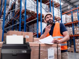 Epson mantiene vigente la impresión por matriz de puntos en operaciones críticas Composición visual que muestra en primer plano una impresora de matriz de puntos Epson de color oscuro con papel continuo instalado. En el fondo, desenfocado para dar profundidad, se observa a un trabajador de logística con casco blanco y chaleco de seguridad naranja, revisando documentos físicos y comunicándose por radio frente a estantes industriales llenos de cajas.