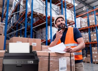 Epson mantiene vigente la impresión por matriz de puntos en operaciones críticas Composición visual que muestra en primer plano una impresora de matriz de puntos Epson de color oscuro con papel continuo instalado. En el fondo, desenfocado para dar profundidad, se observa a un trabajador de logística con casco blanco y chaleco de seguridad naranja, revisando documentos físicos y comunicándose por radio frente a estantes industriales llenos de cajas.