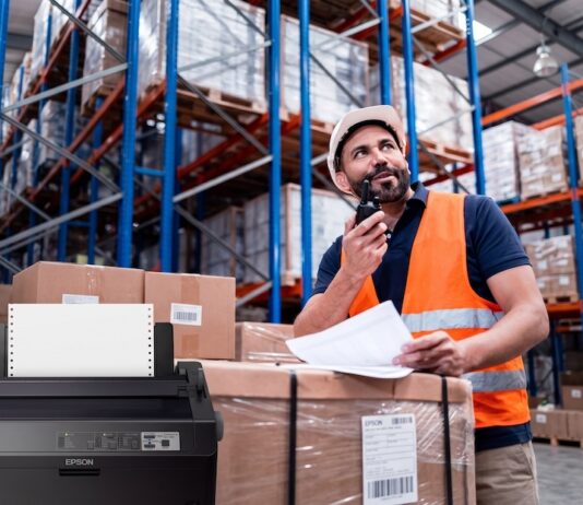 Epson mantiene vigente la impresión por matriz de puntos en operaciones críticas Composición visual que muestra en primer plano una impresora de matriz de puntos Epson de color oscuro con papel continuo instalado. En el fondo, desenfocado para dar profundidad, se observa a un trabajador de logística con casco blanco y chaleco de seguridad naranja, revisando documentos físicos y comunicándose por radio frente a estantes industriales llenos de cajas.