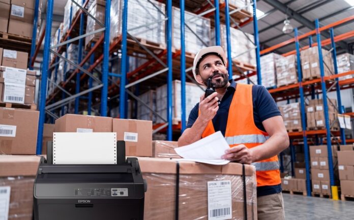 Confiabilidad y trazabilidad ininterrumpida en entornos operativos críticos Composición visual que muestra en primer plano una impresora de matriz de puntos Epson de color oscuro con papel continuo instalado. En el fondo, desenfocado para dar profundidad, se observa a un trabajador de logística con casco blanco y chaleco de seguridad naranja, revisando documentos físicos y comunicándose por radio frente a estantes industriales llenos de cajas.