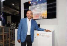 CeleriTech impulsa una nueva etapa para la inteligencia artificial empresarial Fotografía de Gonzalo Bretón, socio cofundador de CeleriTech, sonriendo a la cámara mientras apoya su mano izquierda sobre un módulo blanco con el logotipo naranja y gris de su empresa. Viste un saco azul claro y camisa blanca sin corbata, y se encuentra de pie frente a una gran pantalla que proyecta una presentación corporativa con los logotipos de CeleriTech y SAP.