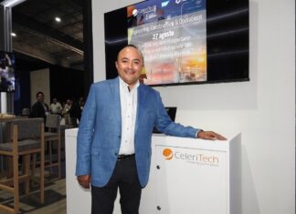CeleriTech impulsa una nueva etapa para la inteligencia artificial empresarial Fotografía de Gonzalo Bretón, socio cofundador de CeleriTech, sonriendo a la cámara mientras apoya su mano izquierda sobre un módulo blanco con el logotipo naranja y gris de su empresa. Viste un saco azul claro y camisa blanca sin corbata, y se encuentra de pie frente a una gran pantalla que proyecta una presentación corporativa con los logotipos de CeleriTech y SAP.