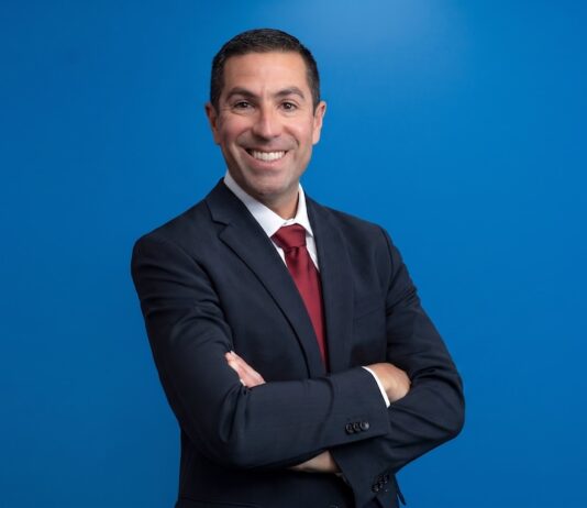 Philips: Ampliar el acceso a la salud exige una nueva forma de innovar Fotografía de medio cuerpo de Negal Ciliberto, posando sonriente y con los brazos cruzados mirando directamente a la cámara. Viste un traje oscuro formal, camisa blanca y corbata roja, resaltando sobre un fondo azul sólido y uniforme.