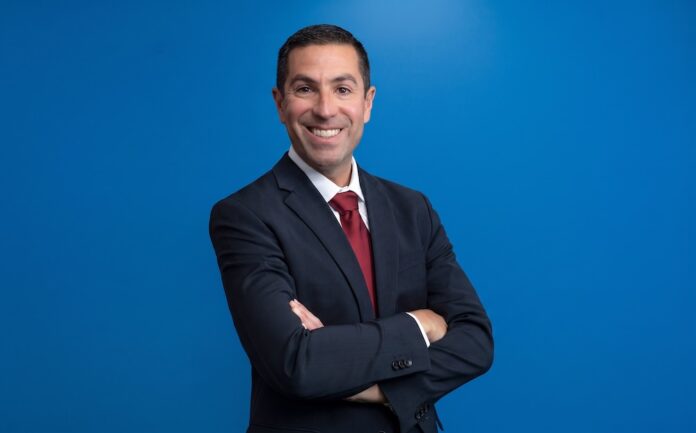 Ecosistemas conectados. El futuro de la atención médica oportuna Fotografía de medio cuerpo de Negal Ciliberto, posando sonriente y con los brazos cruzados mirando directamente a la cámara. Viste un traje oscuro formal, camisa blanca y corbata roja, resaltando sobre un fondo azul sólido y uniforme.
