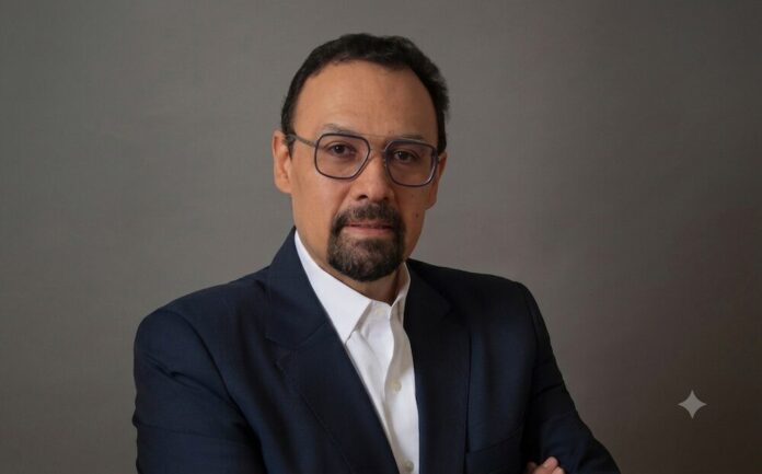 El desafío de la velocidad en la ciberseguridad moderna Fotografía de retrato de Jorge López, un hombre con barba candado, gafas de montura fina y traje oscuro con camisa blanca, mirando directamente a la cámara sobre un fondo gris neutro.